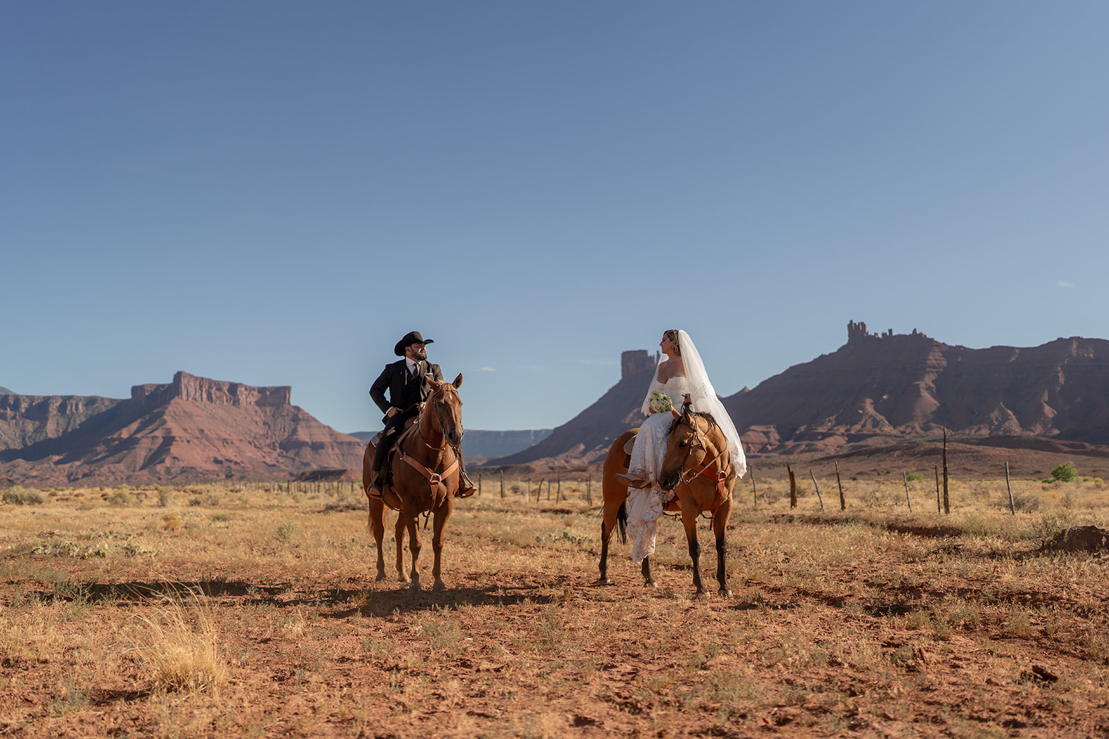 The couple is looking confidently forward in a wide desert setting with prominent red rock formations in the background. Moab elopement with Red Cliffe Horses.