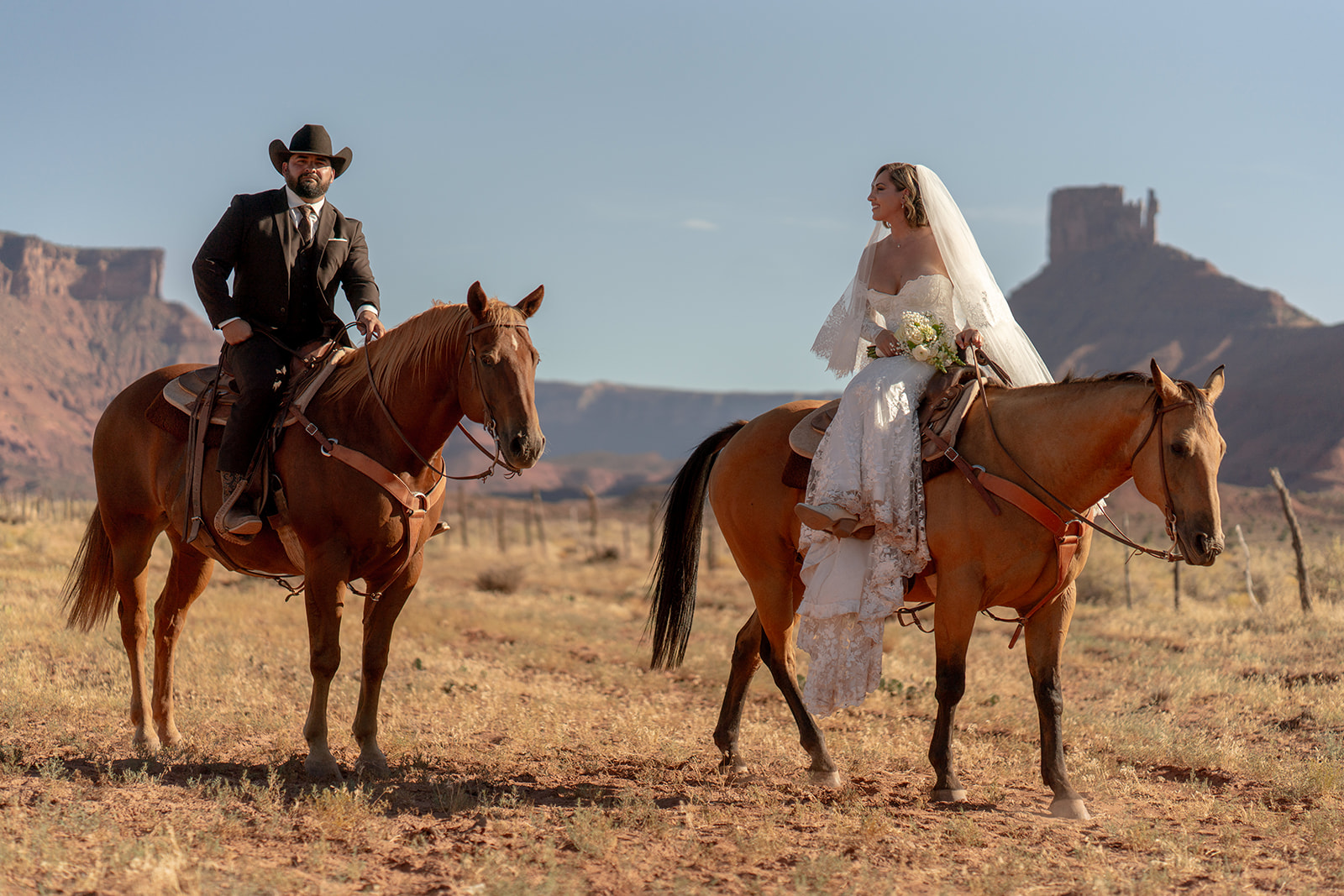 The couple is looking confidently forward in a wide desert setting with prominent red rock formations in the background. Moab elopement with Red Cliffe Horses.