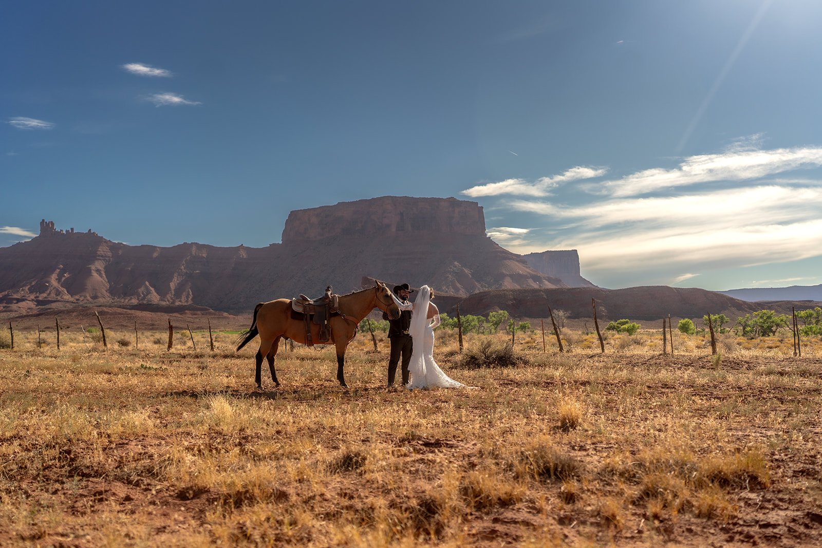Bride and groom embracing in their wedding attire next to a saddled horse in a dry, open field, with massive red rock buttes and the La Sal Mountains in the background. Moab elopement at golden hour.