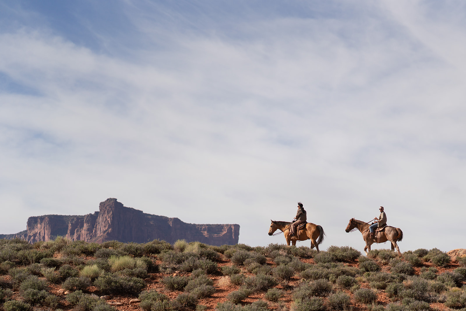 A wide shot of two riders on horseback cresting a scrubby, red desert hill, with a striking flat-topped red rock mesa dominating the horizon beneath a cloudy blue sky