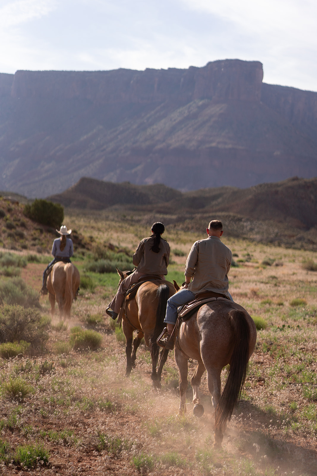 Three people on horseback riding away from the camera on a dusty trail.