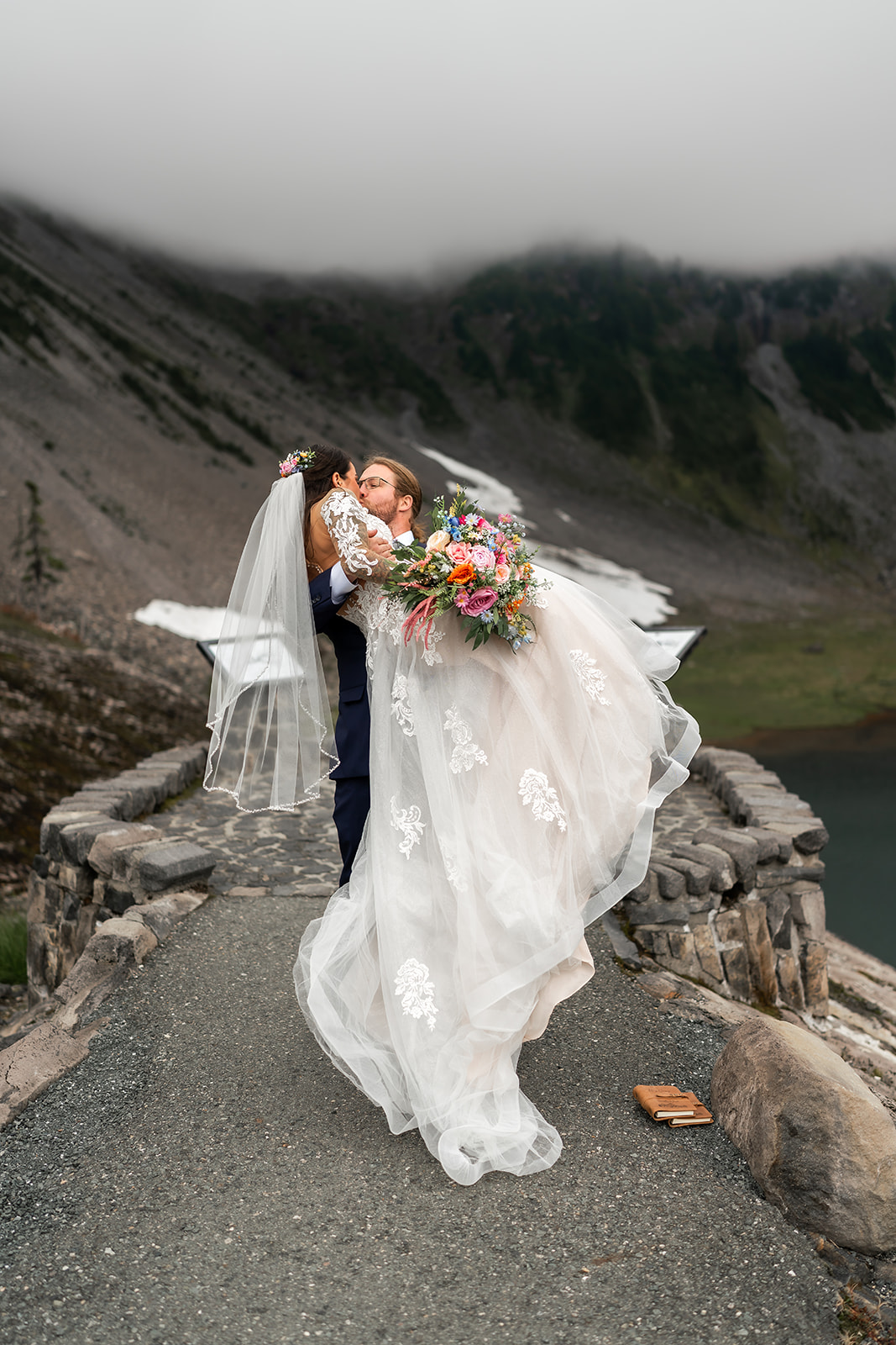 A groom in a navy suit lifts and kisses his bride, who is holding a colorful bouquet
