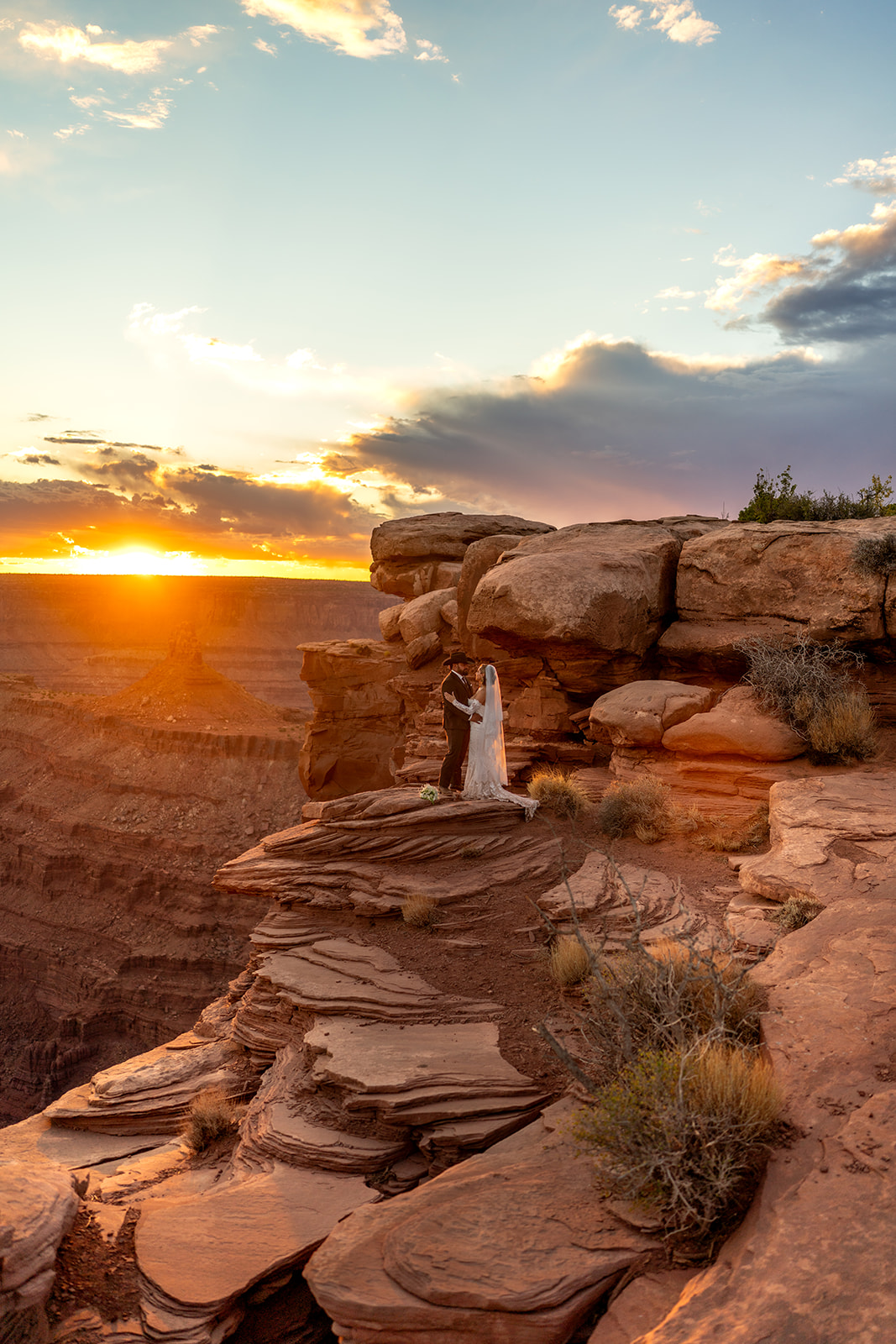 A wide shot of a couple standing on a breathtaking cliff overlooking a canyon at sunset