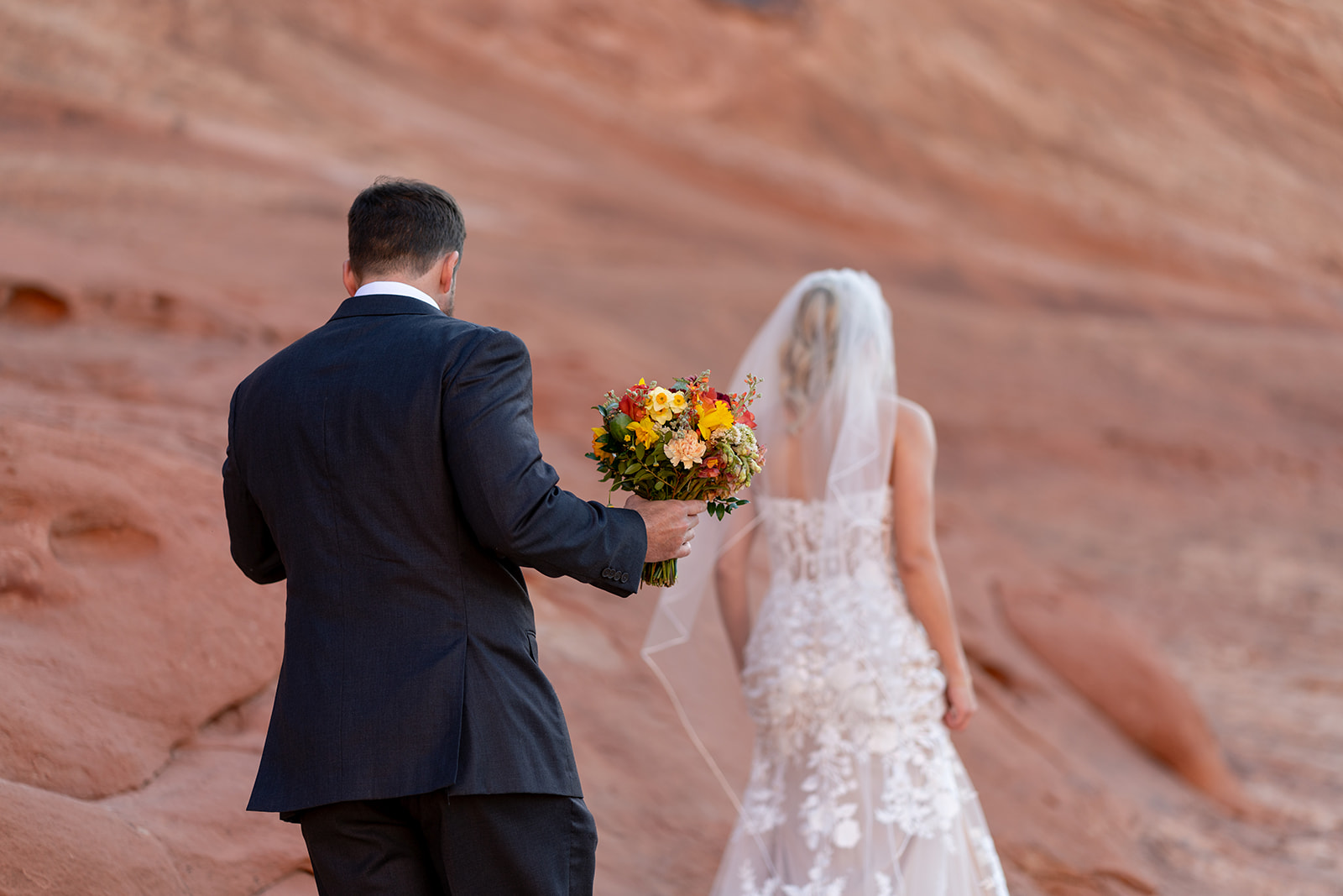 Rear view of elopement couple with the groom holding the custom wildflower-style bouquet by Tangled Sage Event Artistry in the Moab red rock desert.