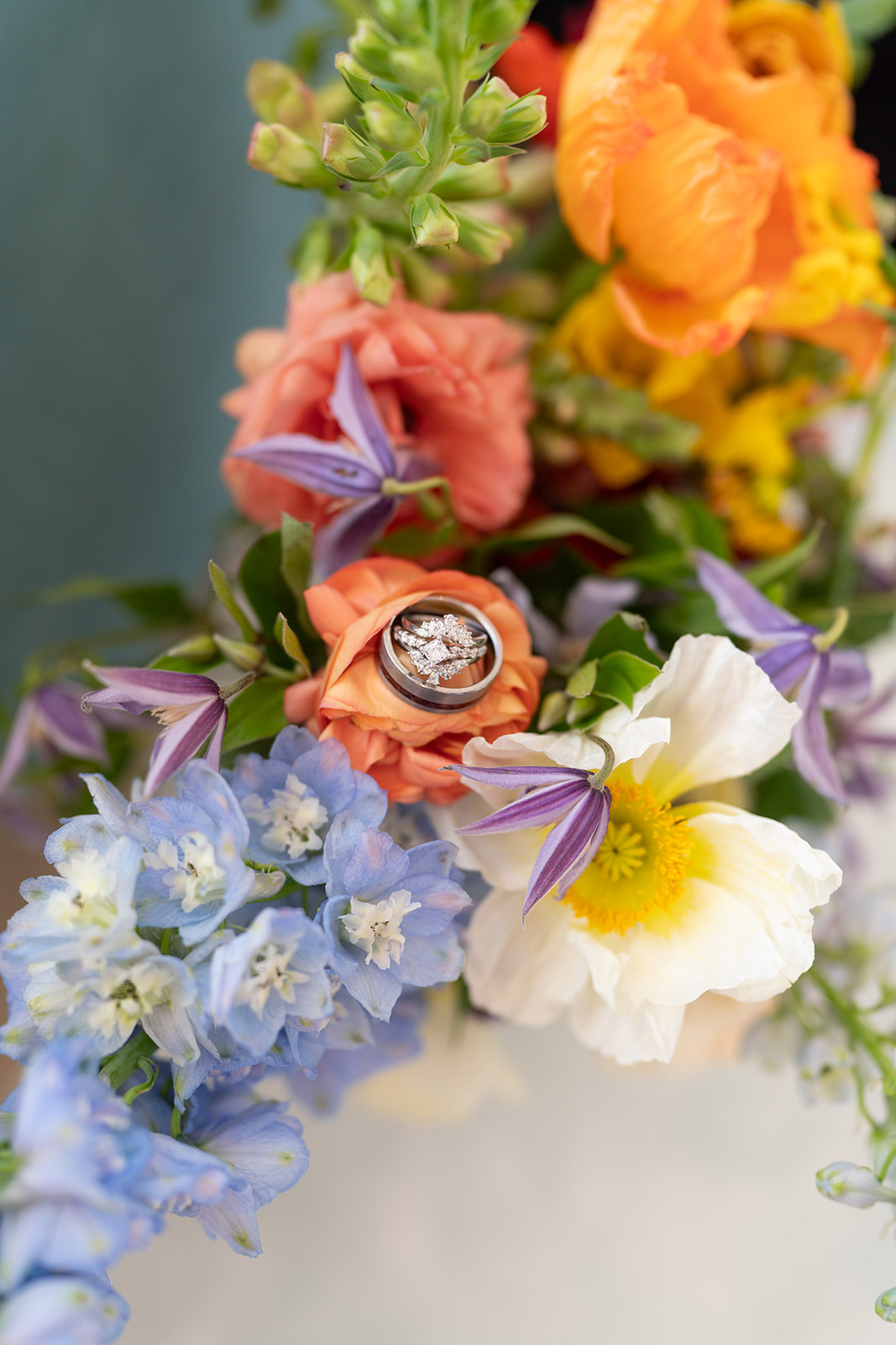 Close-up of wedding rings nestled in a colorful spring bouquet featuring blue, purple, orange, and white flowers, designed by Tangled Sage Event Artistry