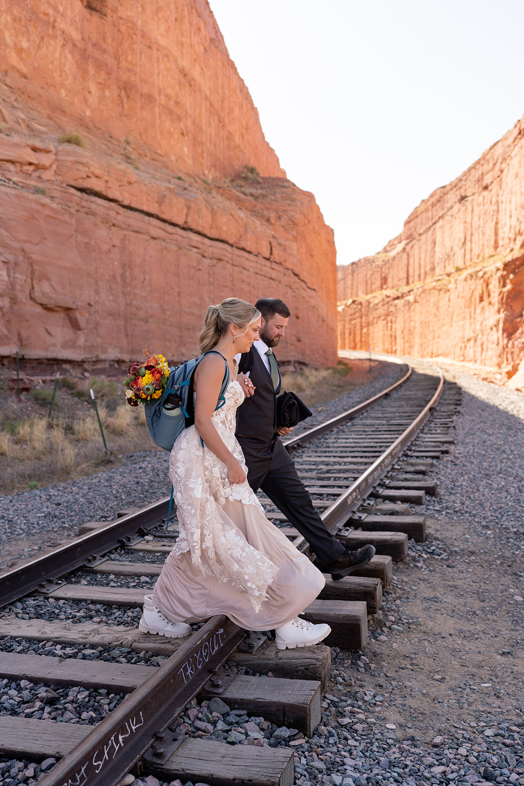 Adventurous elopement couple walking on train tracks in Moab canyon; the bride carries her vibrant bouquet, created by Tangled Sage Event Artistry, in a blue backpack