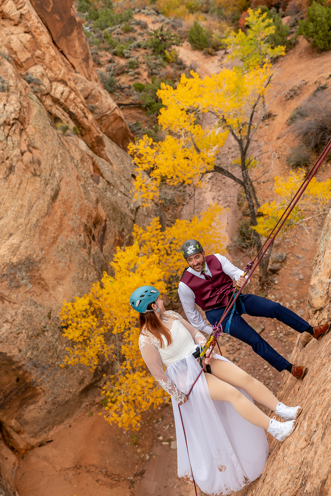Close-up of a smiling couple in wedding attire and helmets rappelling down a canyon wall, with bright yellow aspen trees in the background
