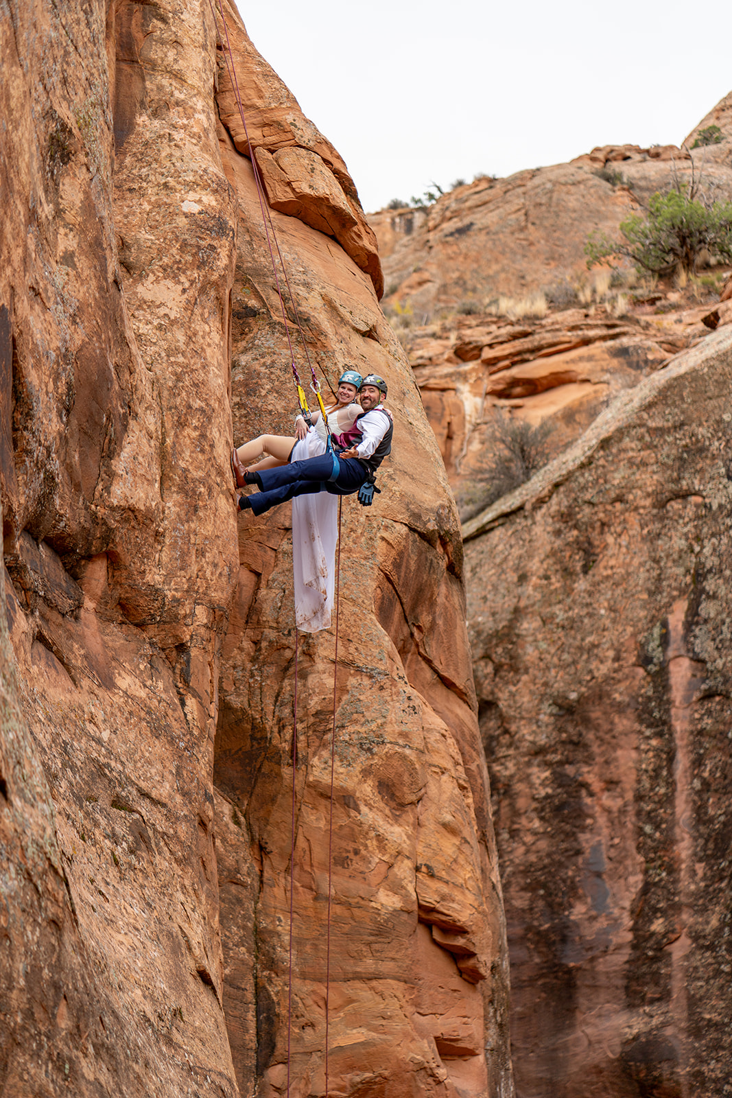 A newlywed couple in wedding attire rappels down a steep, orange-colored rock face in Moab, Utah