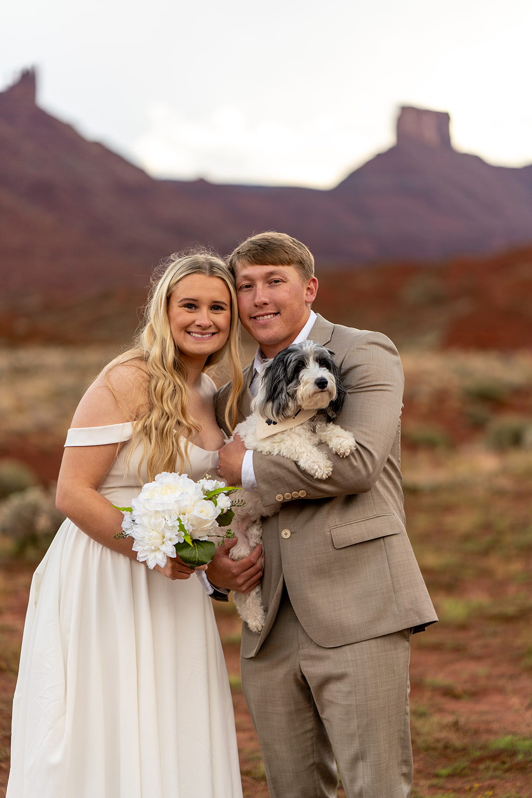 Smiling elopement couple posing with their small black and white dog against the red rock desert landscape of Castle Valley Moab Utah