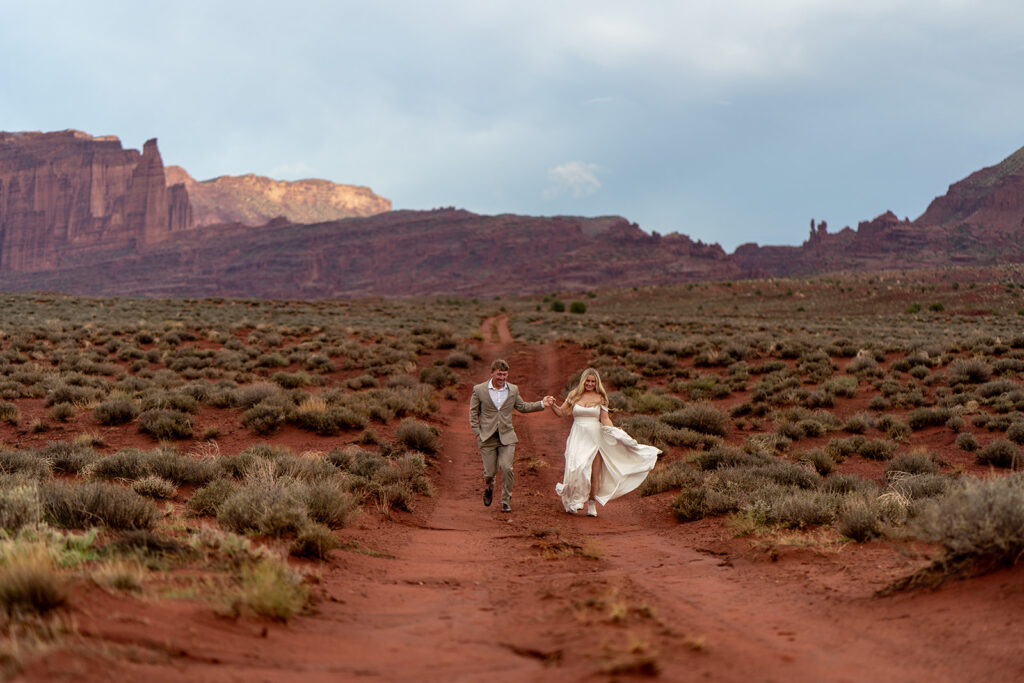 Newlyweds celebrating post-elopement joy by running hand-in-hand down a red dirt road in the Moab, Utah desert