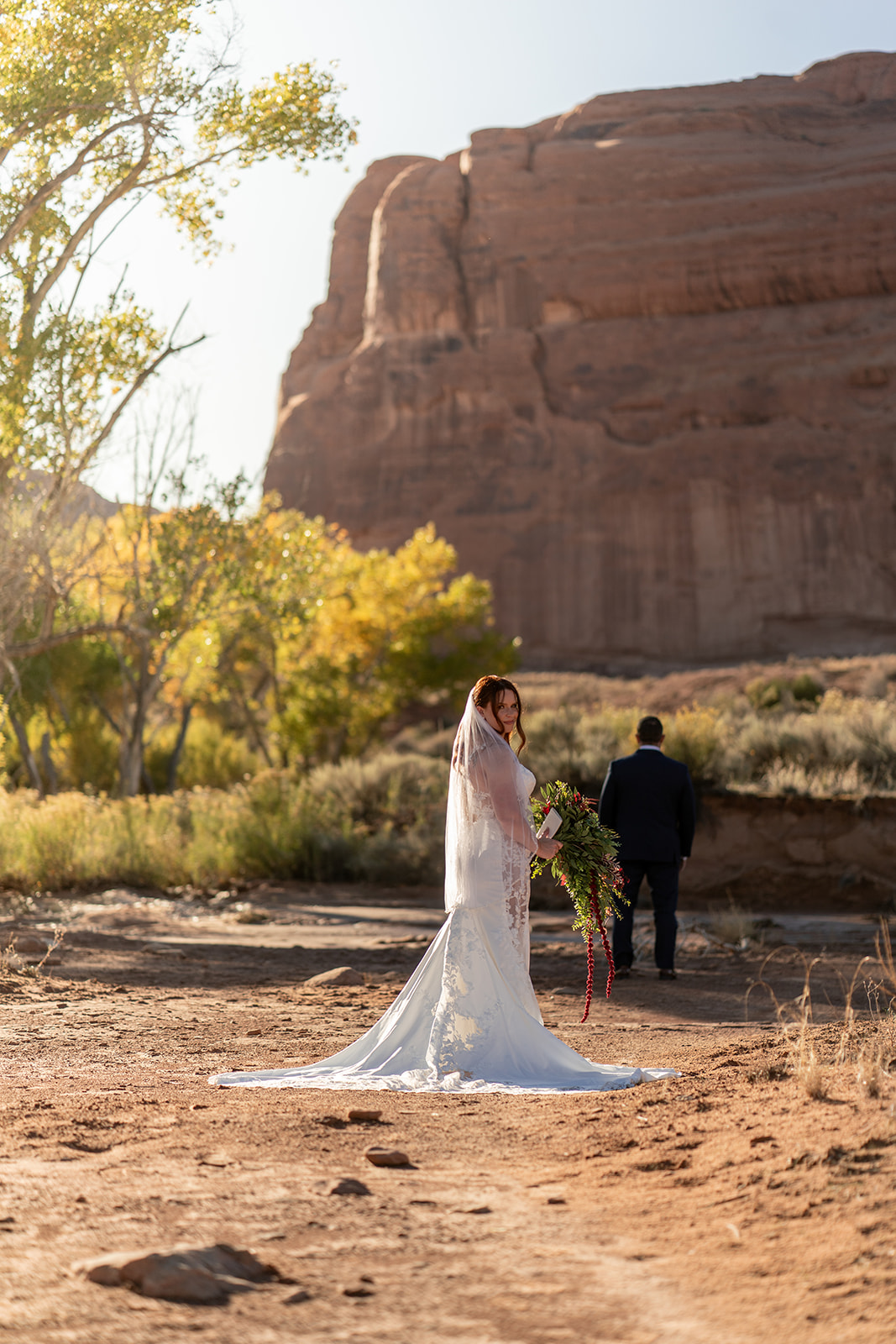 A bride in a lace wedding dress with a trailing veil and a large bouquet stands in a dry riverbed, looking over her shoulder. The groom walks away toward a massive, rounded sandstone cliff in the background