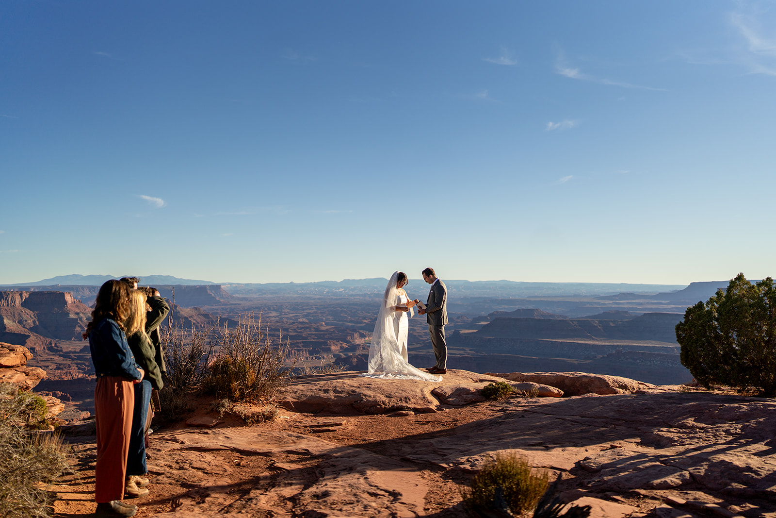 A couple in wedding attire exchanges vows at a scenic overlook in Moab, Utah, with vast canyons stretching into the distance and two witnesses watching knowing The Cost to Elope in 2025