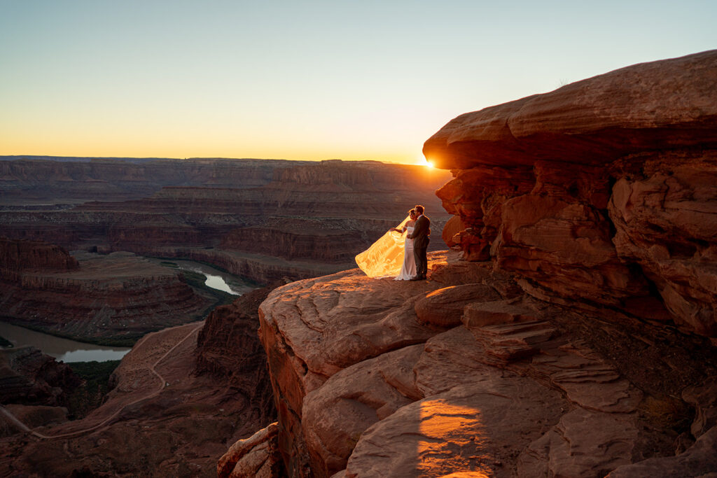 Wide shot of a married couple embracing on a rocky cliff overlooking a deep desert canyon and river at sunrise, with the bride's veil backlit by the sun
