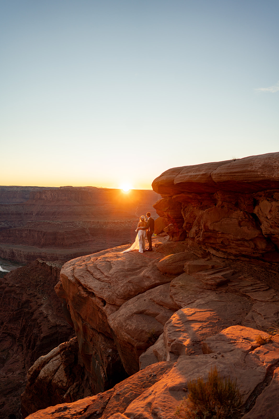A newlywed couple stands on a precipitous red-rock cliff edge overlooking a vast canyon landscape during a golden sunset in Utah.