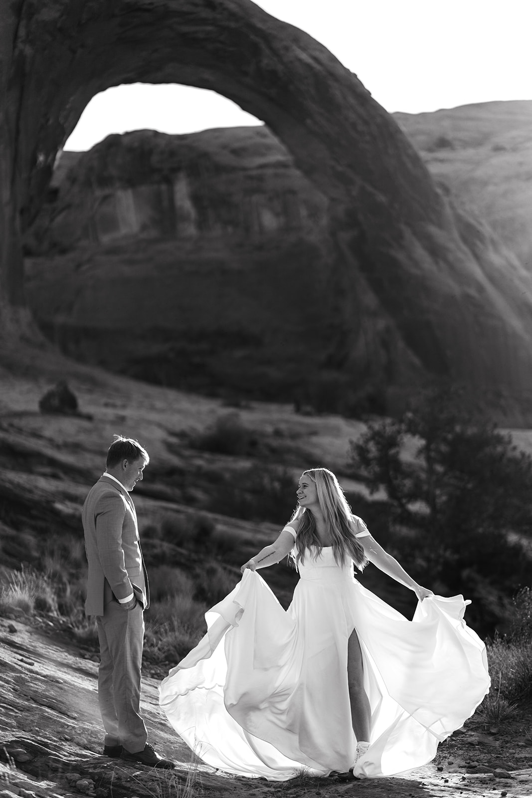 A black and white photograph of an eloping couple in Moab, Utah. The bride is wearing a white dress with a slit, holding the skirt out as if twirling, and smiling at the groom who is looking down at her.